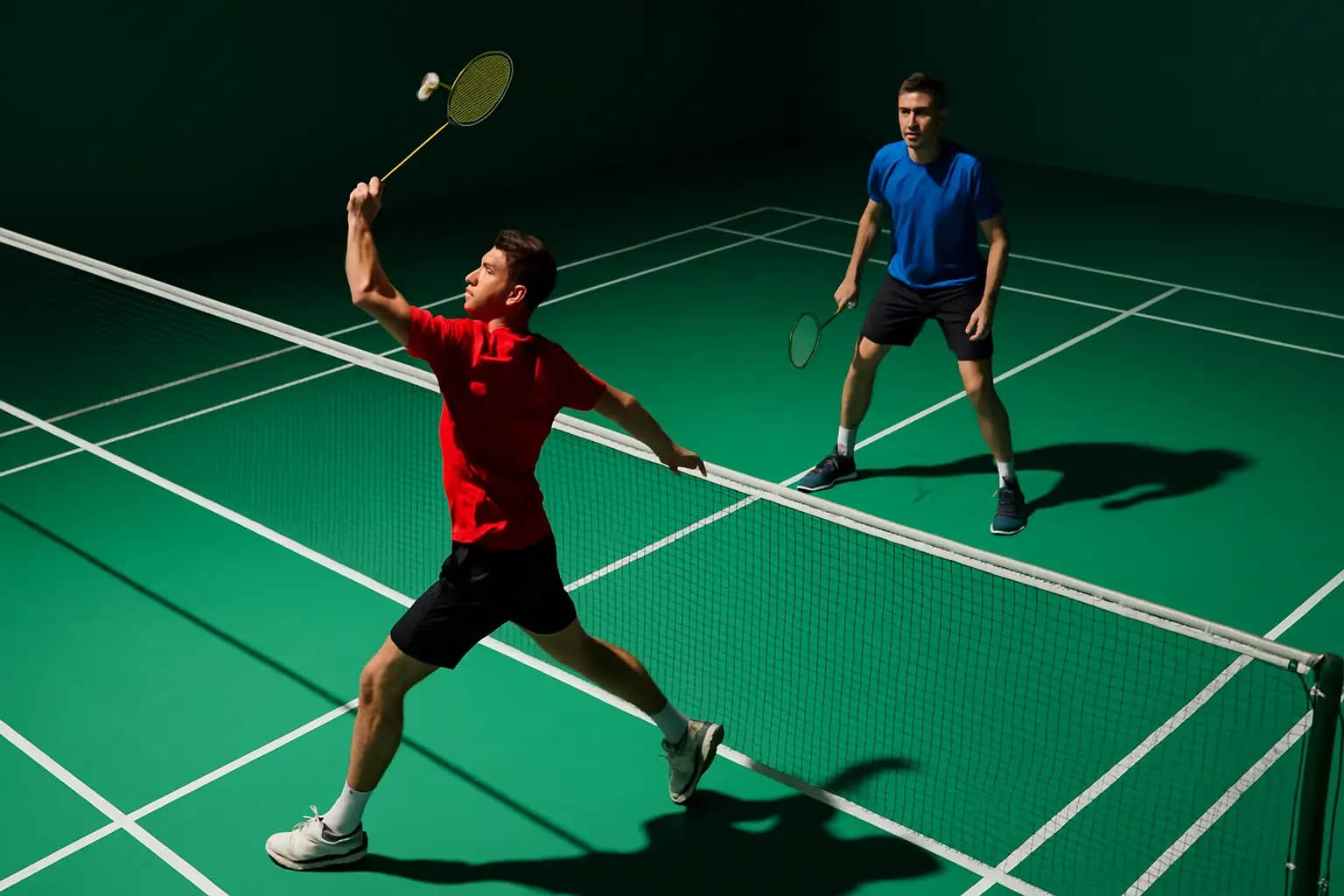 Malaysian badminton doubles players practicing on an indoor court with rackets raised